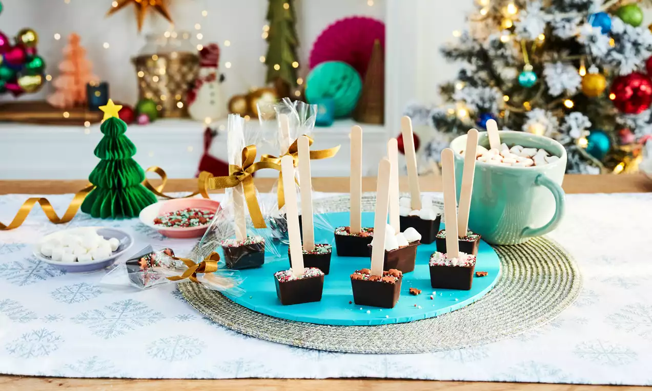 Square blocks of glossy dark chocolate pierced with wooden stirrers, sprinkled with colourful decorations on a turquoise plate