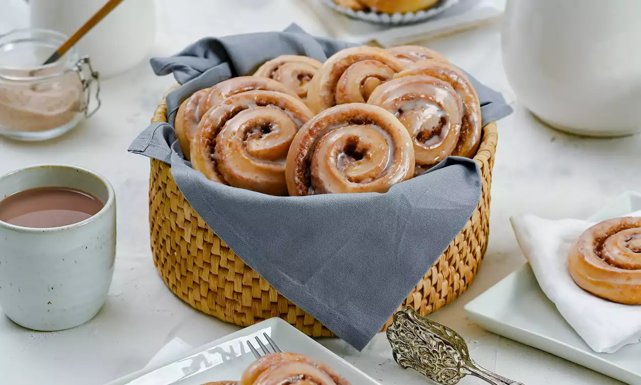 Goldbraune Zimtschnecken mit glänzender Zuckerglasur in einem geflochtenen Korb mit grauem Stofftuch, arrangiert auf hellem Untergrund