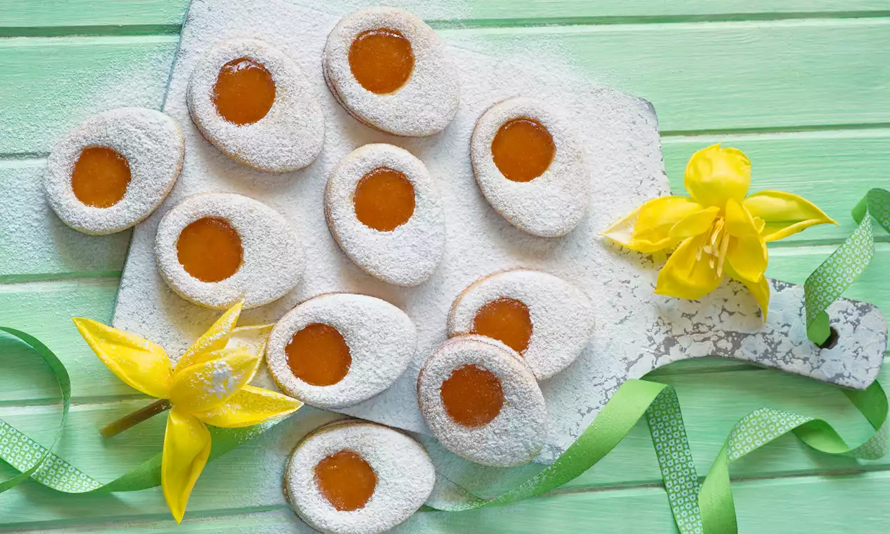 Ovale Linzer Plätzchen mit orangenem Aprikosenkern, bestäubt mit weißem Puderzucker, auf hellgrünem Holzbrett mit gelber Narzisse drapiert