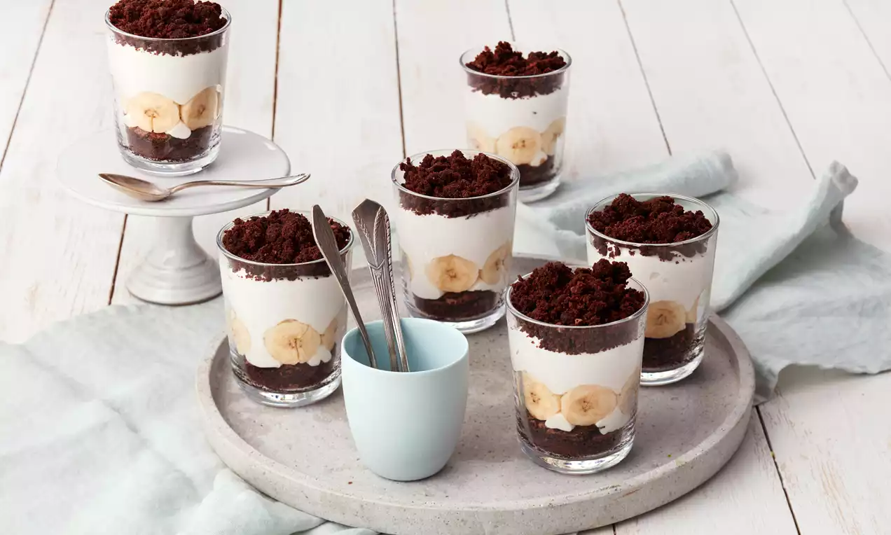 Schichtdesserts im Glas mit hellem Sahnecreme, Bananenscheiben, dunklem Schokokrümelboden und Schokostreuseltopping auf hellem Serviergeschirr 