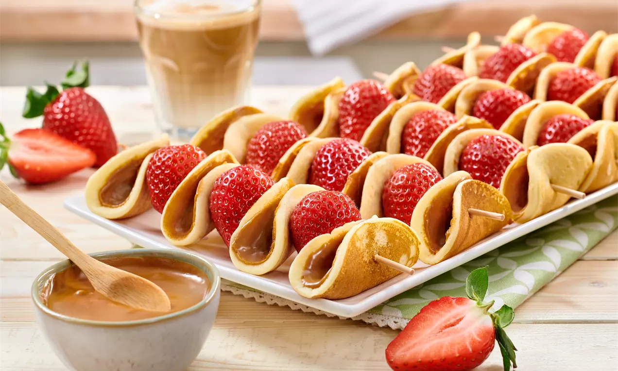 Spieße mit goldgelben Mini-Pfannkuchen und leuchtend roten Erdbeeren, arrangiert auf einem weißen rechteckigen Teller mit Karamellsauce in einer Schale