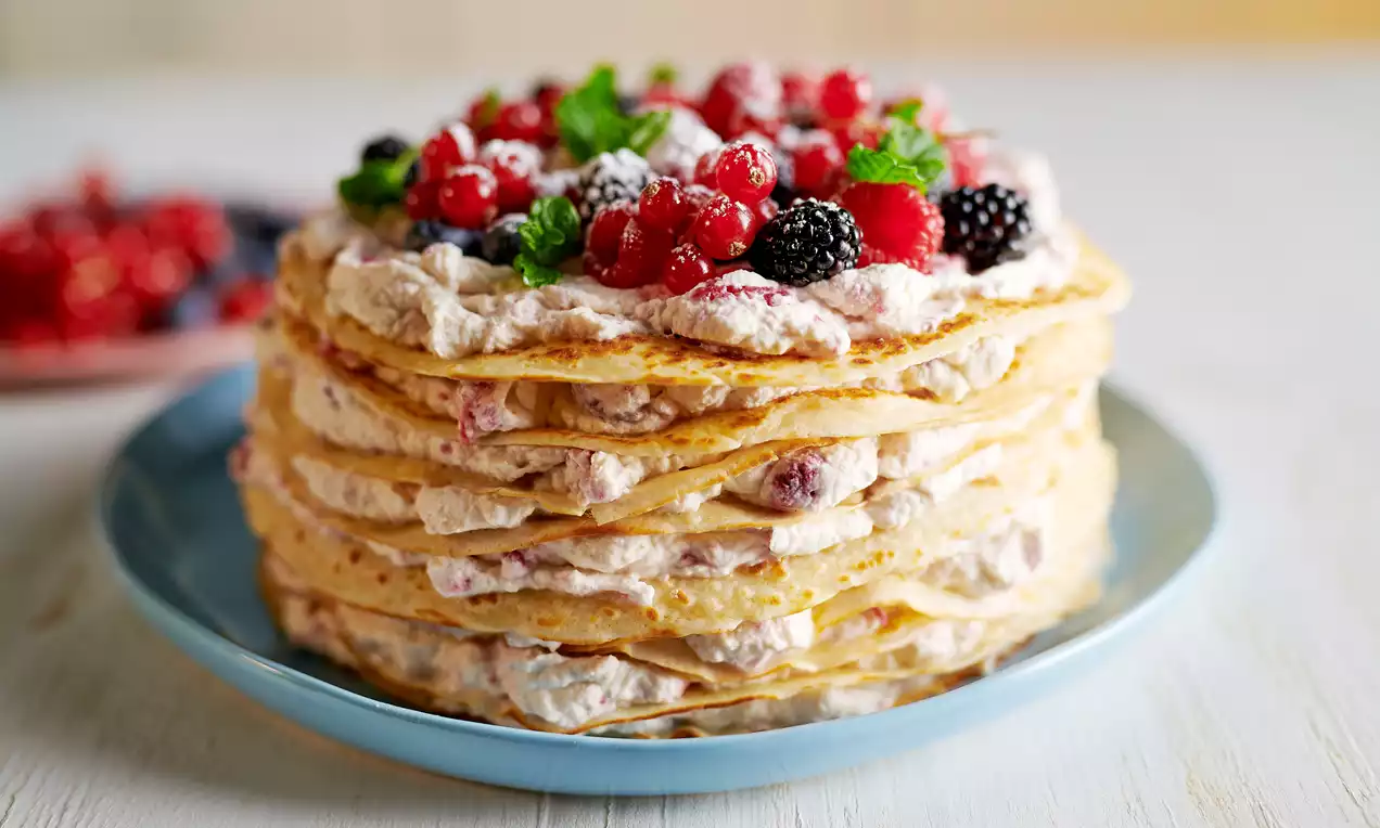 Tarte de crêpes empilées avec des couches de crème fouettée, garnie de fruits rouges, de feuilles de menthe et d’un voile de sucre glace
