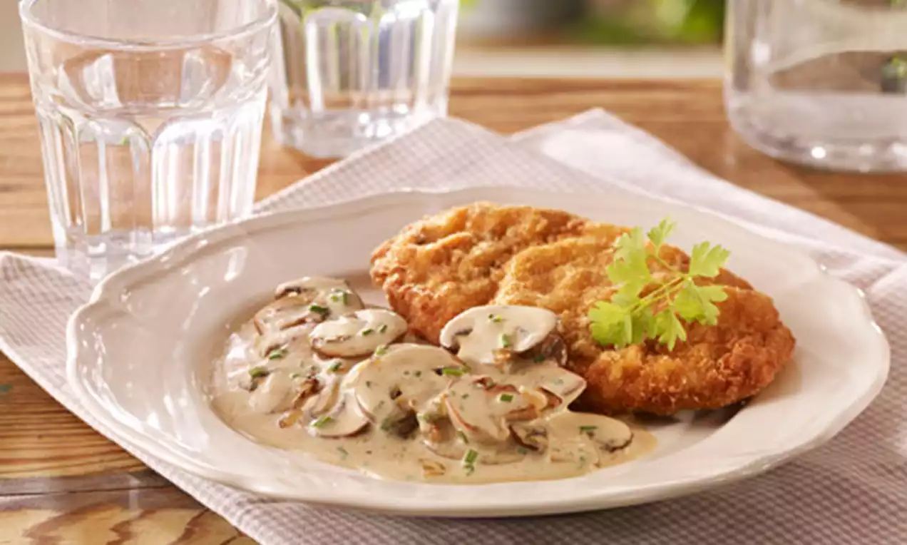 Goldbraunes Schnitzel mit cremefarbener Pilzsauce, garniert mit frischer Petersilie, angerichtet auf weißem Teller mit Holzunterlage und Gläsern
