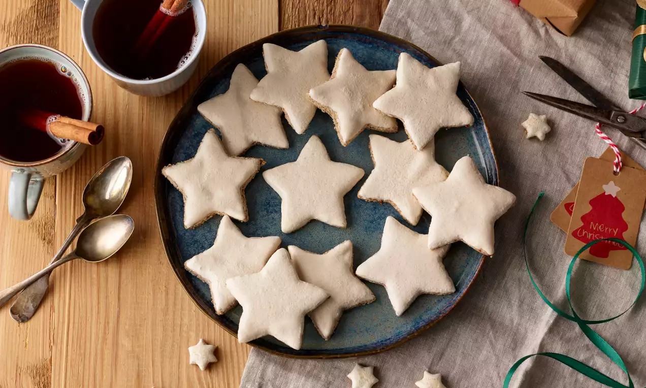 Zimtsterne mit weißem Guss und weicher Textur auf einem runden blauen Teller, arrangiert mit Teetassen, Löffeln und Weihnachtsdekoration