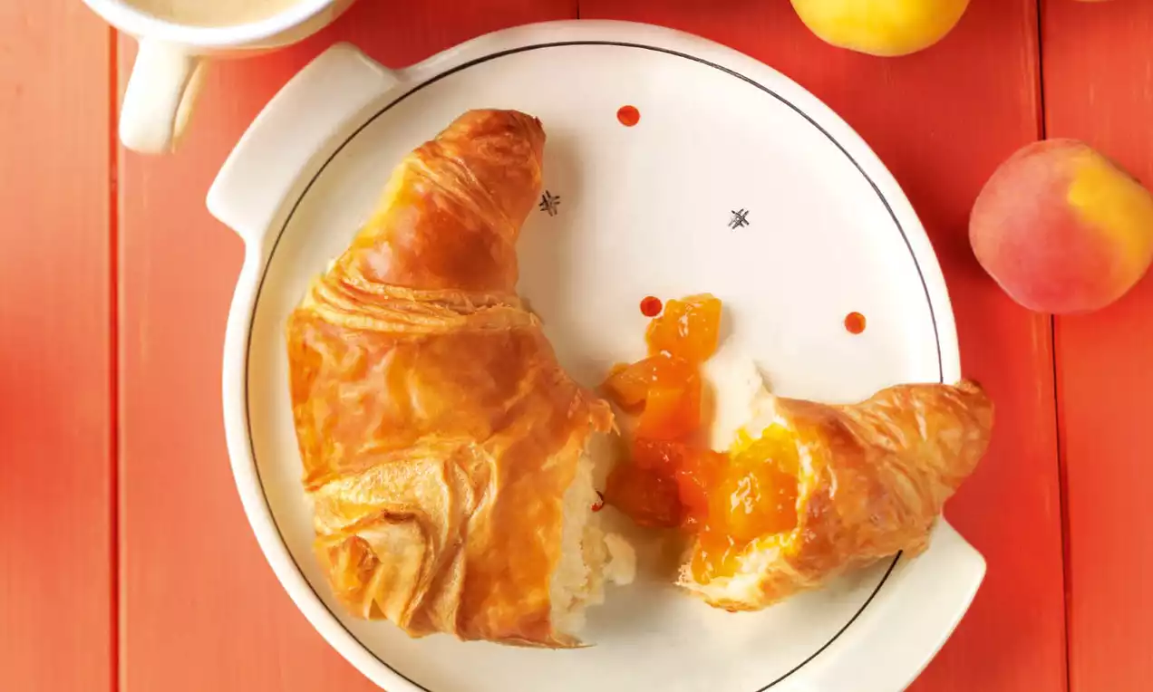 Džem od marelica i manga svijetlonarančaste boje izlazi iz zlatnopečenog kroasana na bijelom tanjiru s crvenim i crnim detaljima