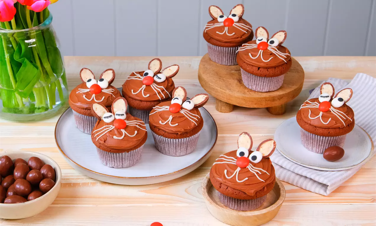 Schokoladenmuffins mit Hasengesichtern aus roten Nasen, weißen Zuckerglasur-Linien und Mandelohren auf Tellern und Holzständern angerichtet