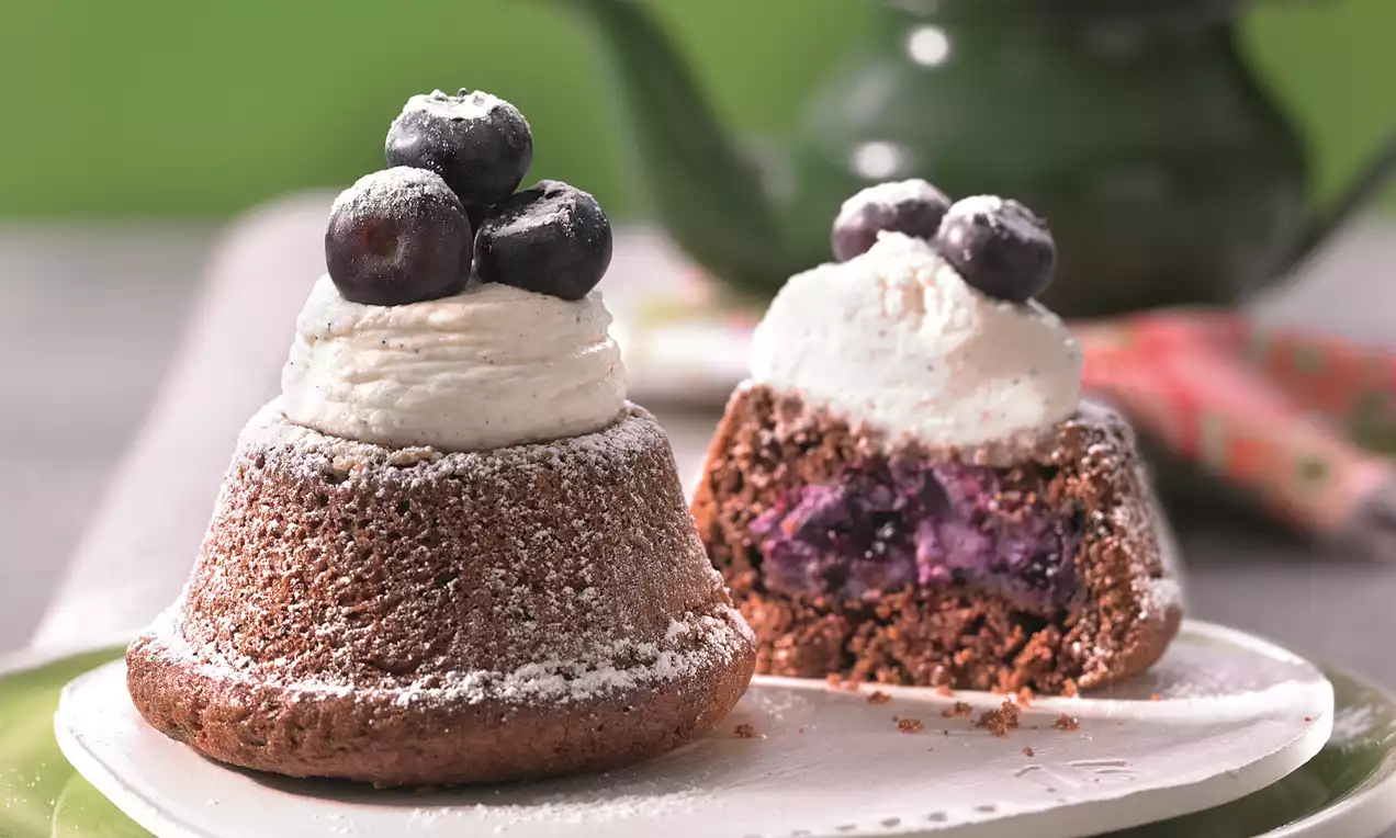 Kolačić smeđe boje s prhkom teksturom ukrašen kremom i svježim borovnicama, unutrašnjost s ljubičastim punjenjem na bijelom tanjuru