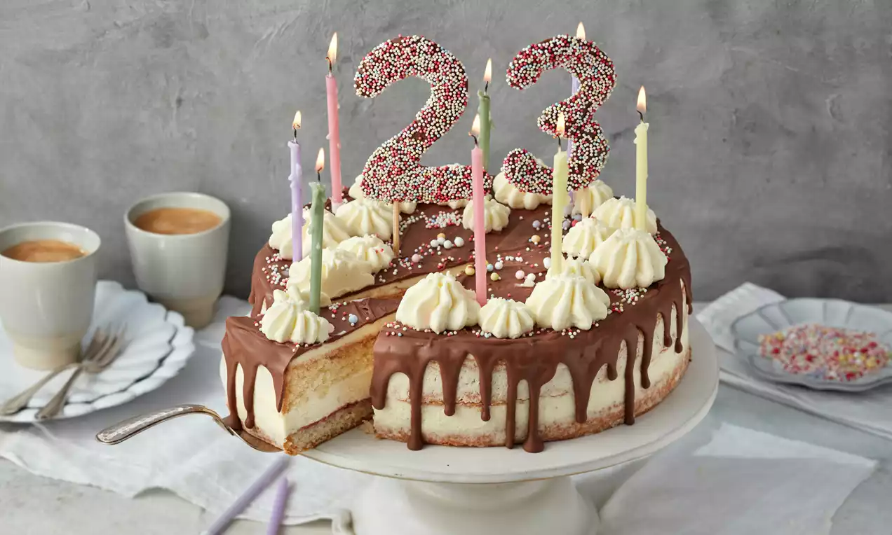 Geburtstagstorte mit Schokoladenglasur, Sahnetupfen, bunter Zuckerstreu und Kerzen, auf weißer Tortenplatte mit angeschnittenem Stück