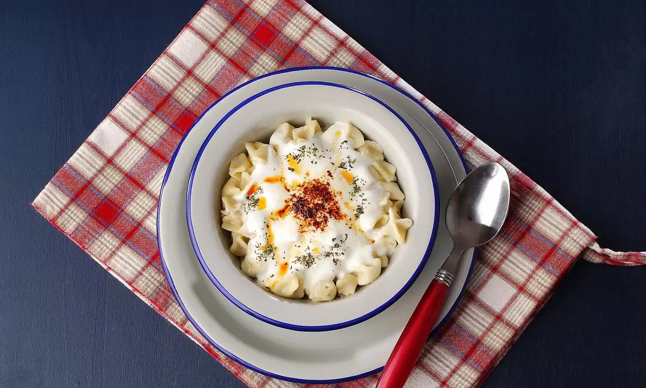 Beyaz seramik kasede sunulan glutensiz mantı, üstünde yoğurt, kızdırılmış tereyağı, toz biber ve kuru nane ile kareli örtü üzerinde.