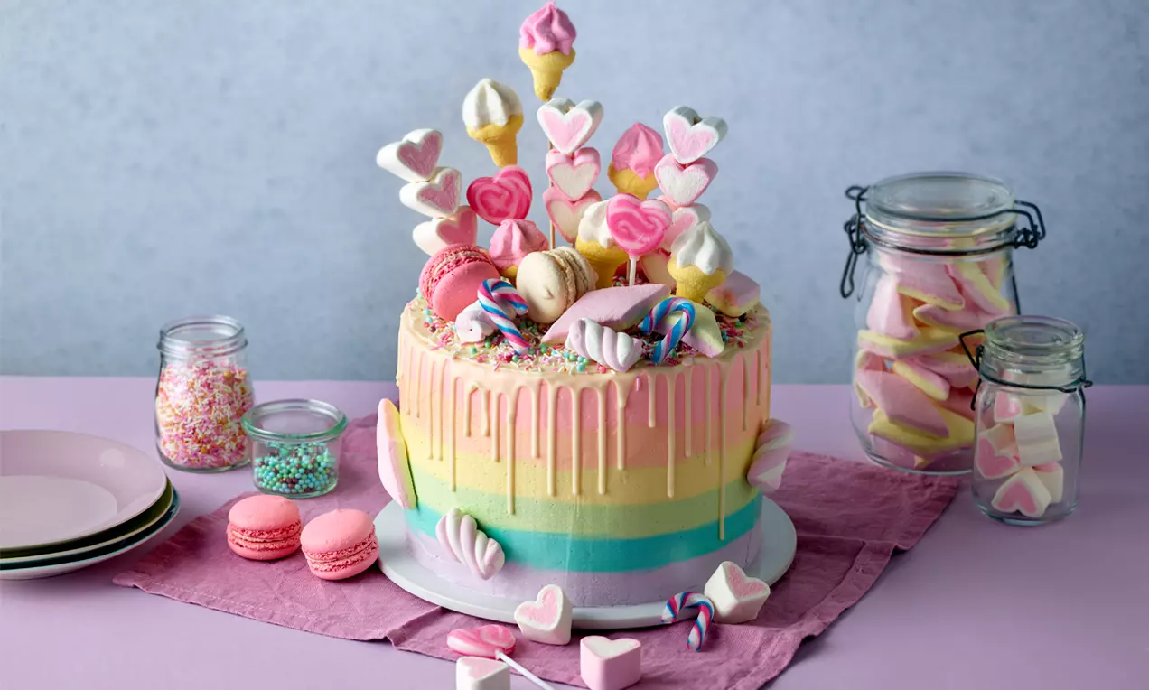 Bunte, gestreifte Torte mit pastellfarbenem Drip, dekoriert mit Macarons, Marshmallows, Zuckerstangen und Herzspießen auf violettem Tuch