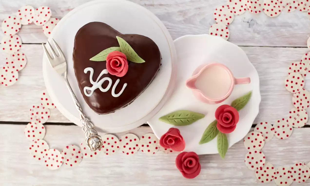 Herzförmiger Mini-Kuchen mit glänzender Schokoglasur, dekoriert mit grünen Blättern, roten Marzipanrosen und weißem Schriftzug auf Teller