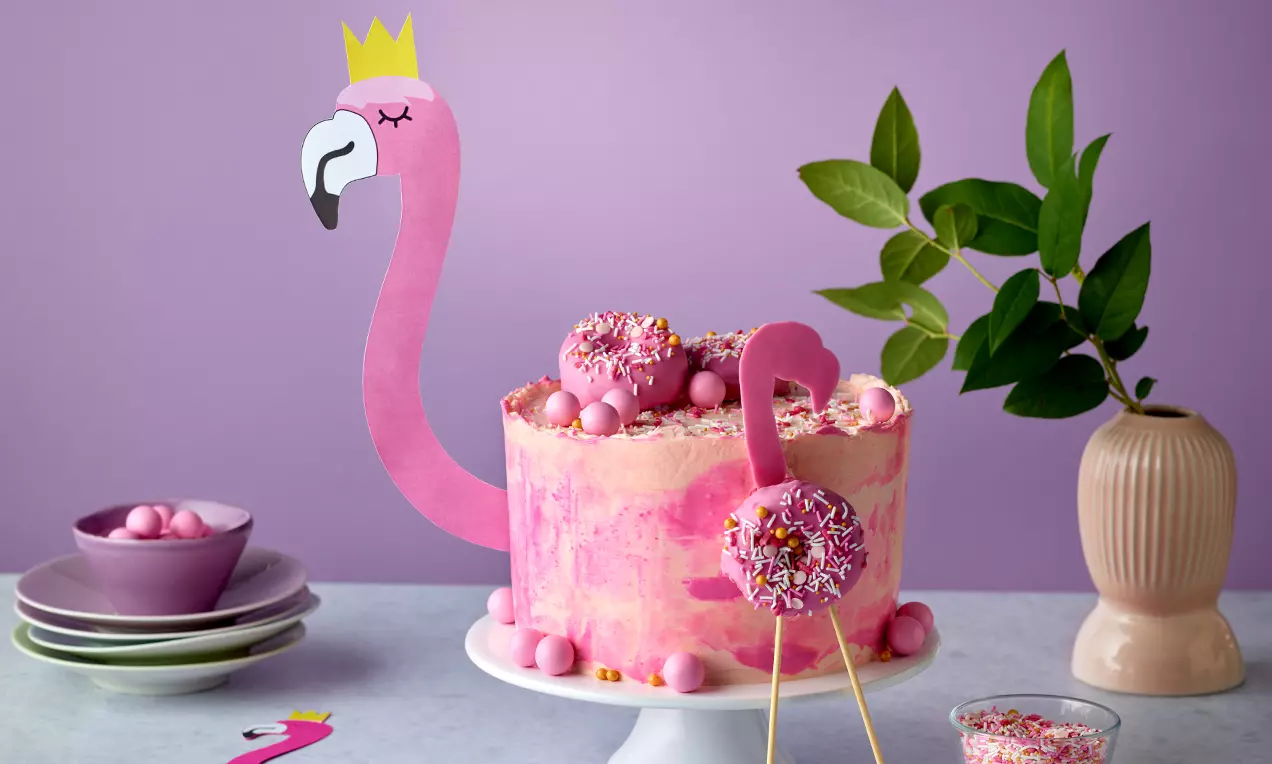 Rosafarbene Flamingo-Torte mit strukturierter Creme, Dekoration aus Donut und Perlen auf weißer Tortenplatte vor violettem Hintergrund