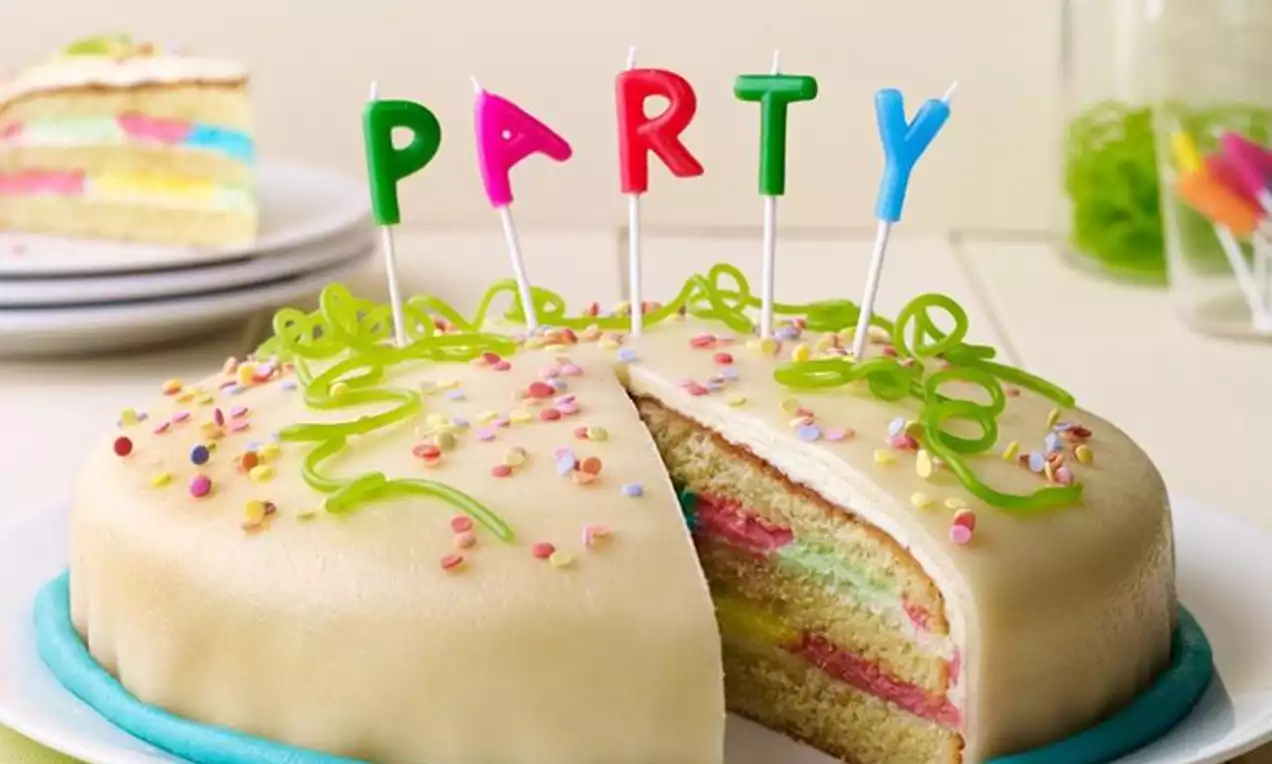 Torta prekrivena svijetložutom glazurom s duginih slojeva biskvita, posuta šarenim mrvicama i ukrašena svijetlozelenim krem trakama 