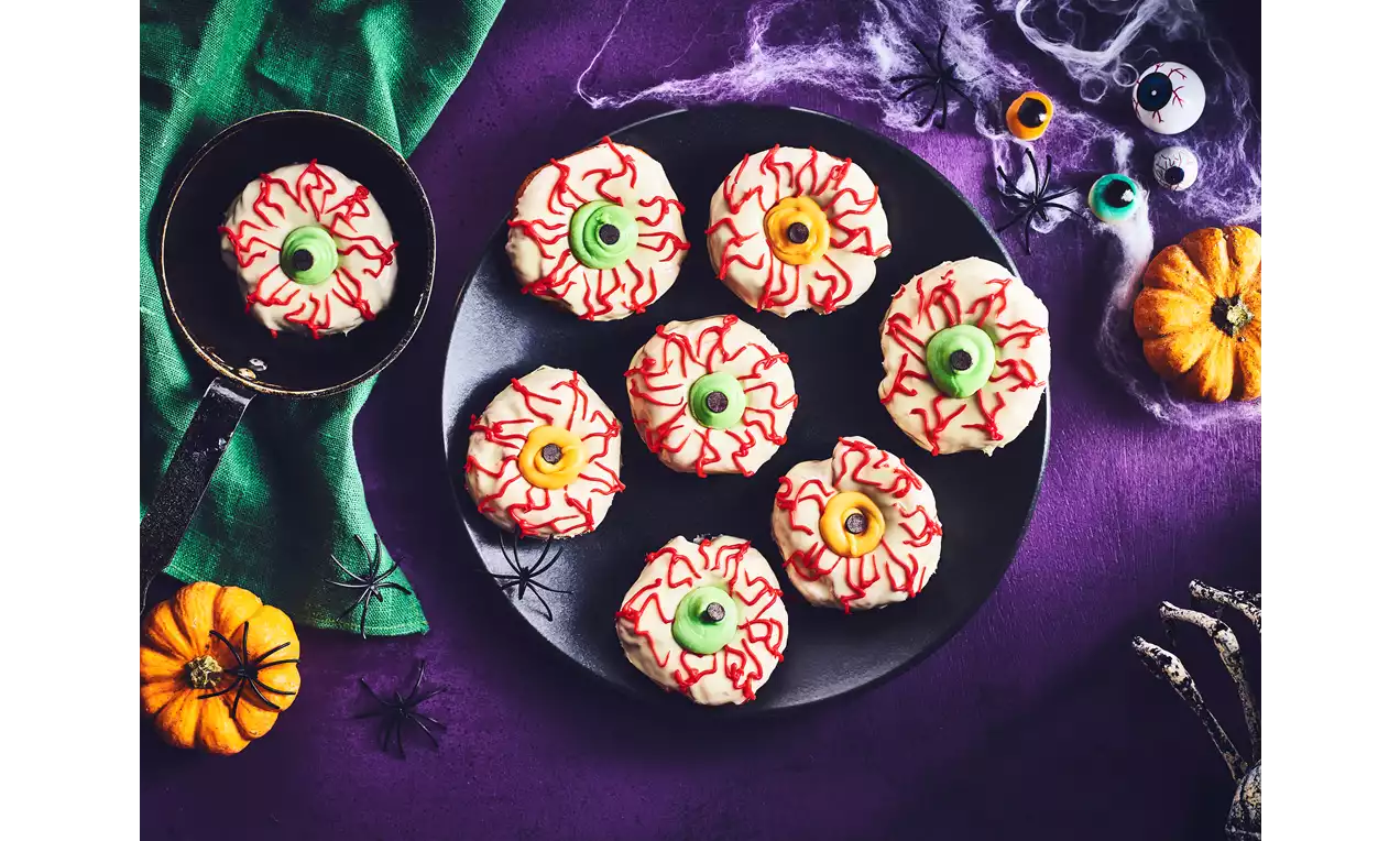 White doughnuts with red icing veins, green and yellow candy eyeballs in the center, arranged on a black plate with pumpkin and spider decor