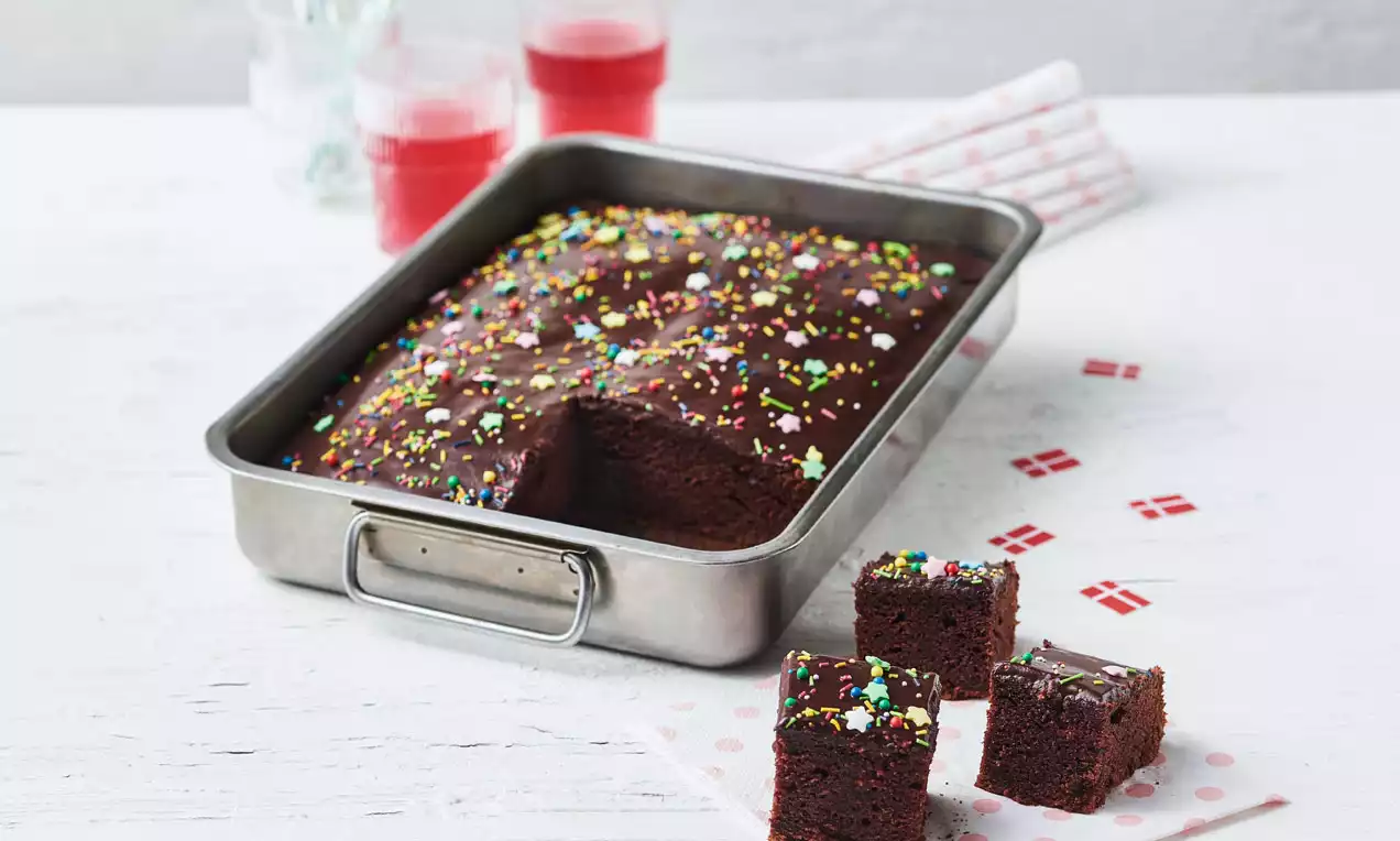 Saftig chokoladekage i en rektangulær metalform, dækket med farverigt krymmel, skåret i firkanter på hvidt bord med flag-servietter