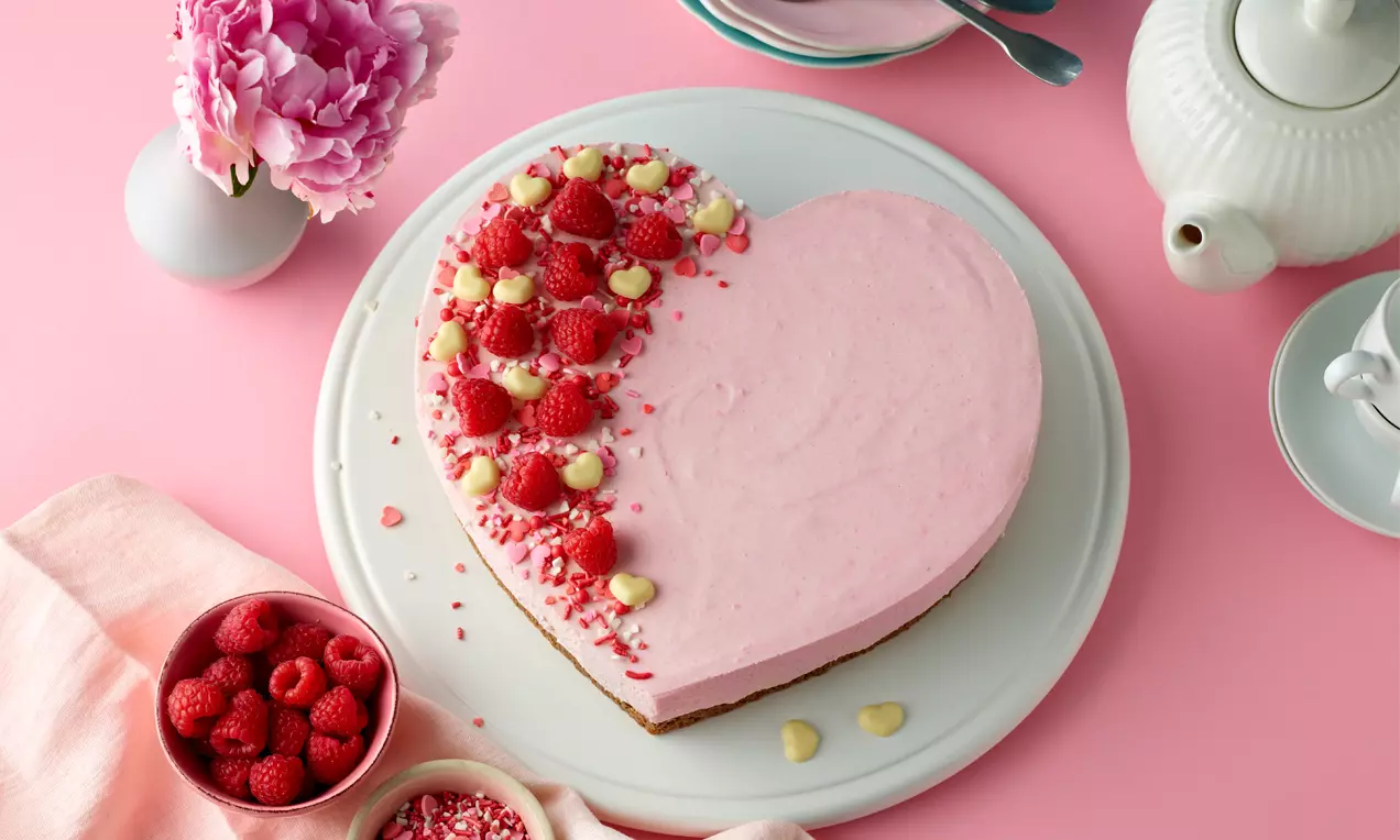 Rosafarbene Himbeermousse-Herztorte auf weißer Platte, mit frischen Himbeeren, weißer Schokoladen-Deko und bunten Streuseln verziert