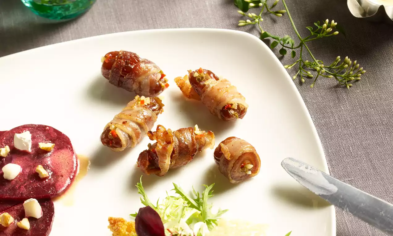 Datteln in knusprigem Speckmantel, goldbraun gebacken, auf weißem Teller, umgeben von Salat, Rote-Bete-Scheiben und filigraner Dekoration
