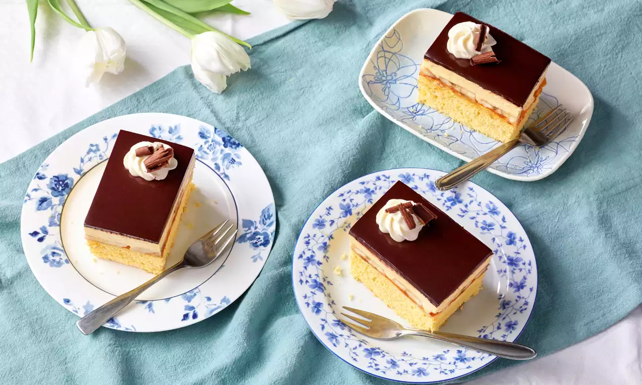 Drei rechteckige Bananenschnitten mit gelbem Biskuit, heller Creme, dunkler Glasur und Sahnetupfen auf floralen Tellern mit Gabel angerichtet