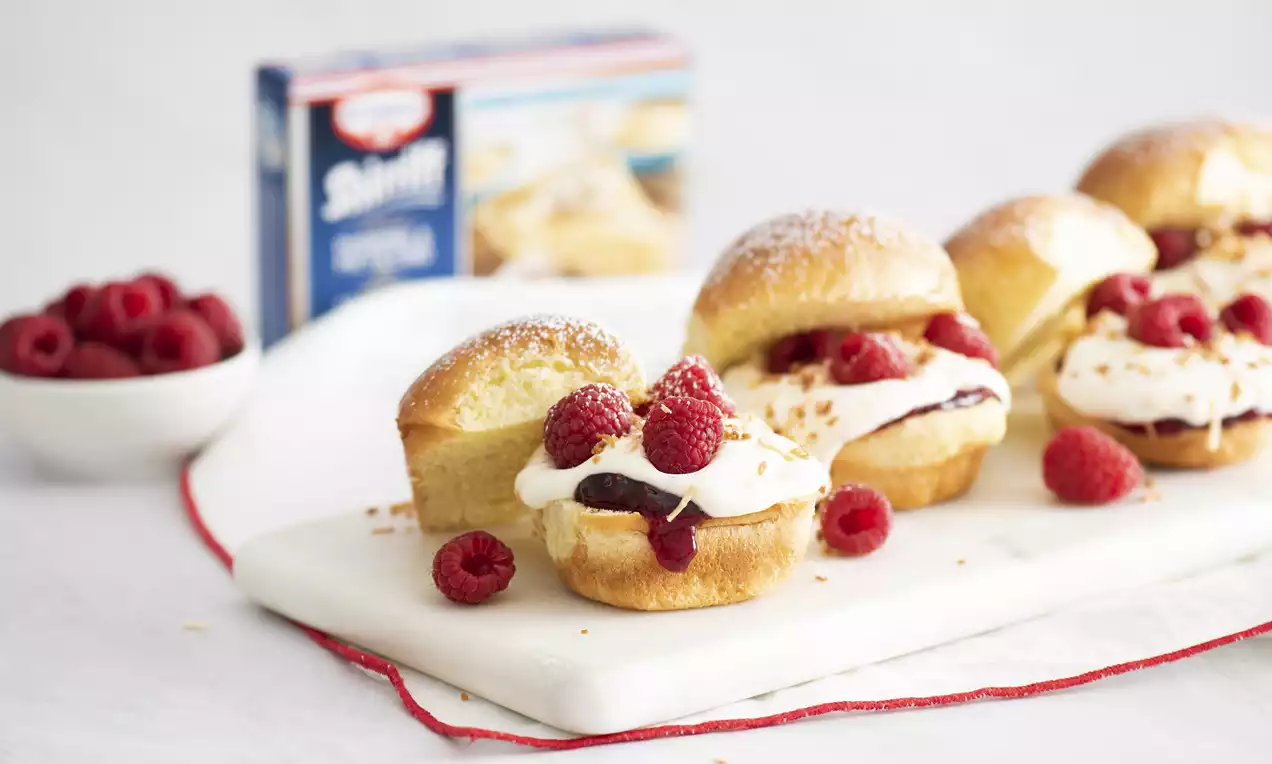 Golden slider buns dusted with powdered sugar, filled with cream, raspberry jam, fresh raspberries, and coconut flakes on a white marble board