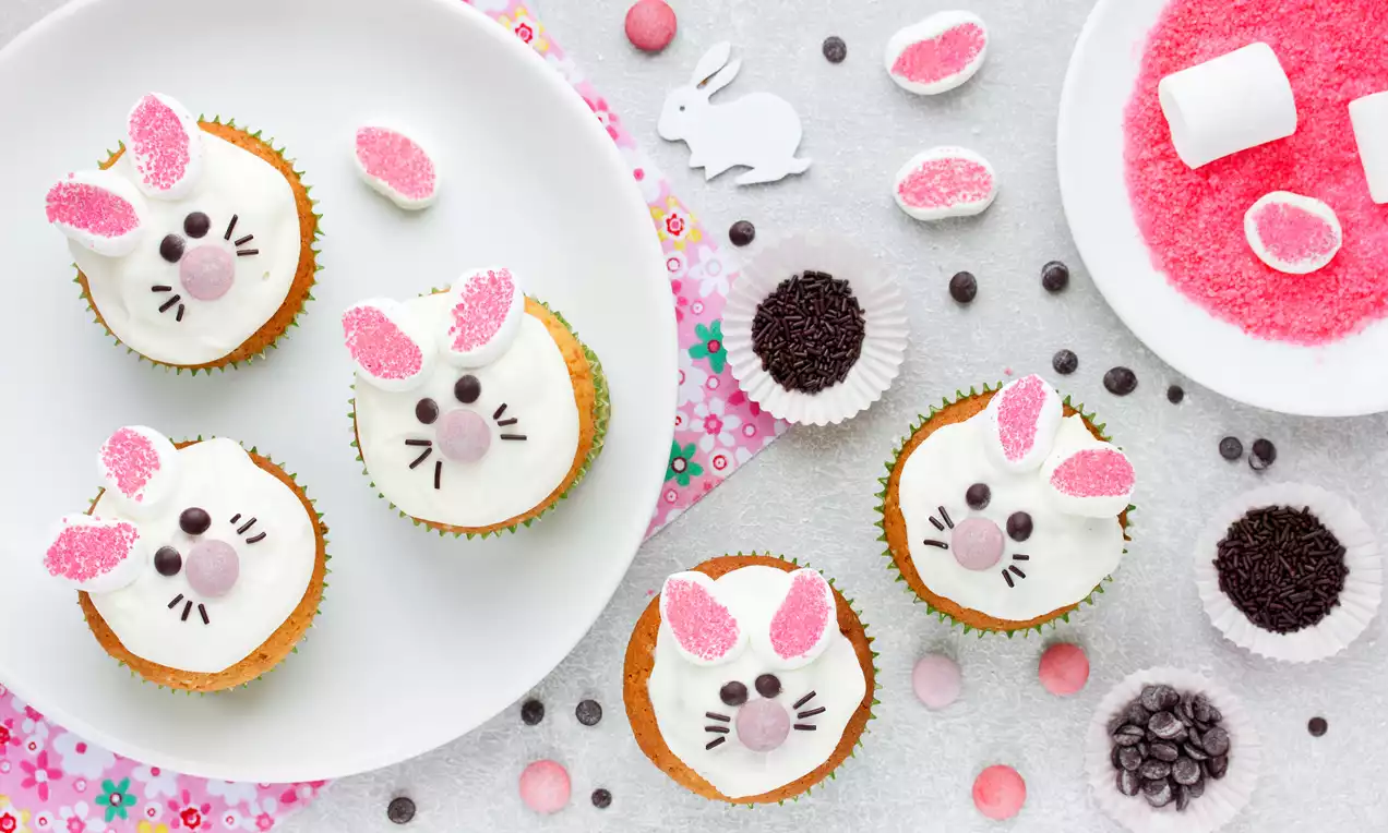 Paascupcakes met witte glazuur, roze-suikeroren, chocoladesnoet en neus, liggend op een wit bord en omlijst door kleurrijke decoraties