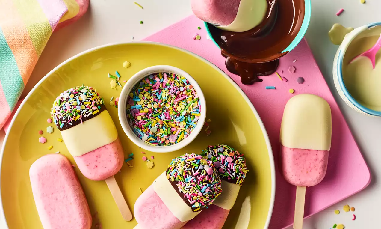 Pink and cream berry chocolate ice lollies with smooth textures, some dipped and sprinkled, arranged on a yellow plate with a sprinkle bowl