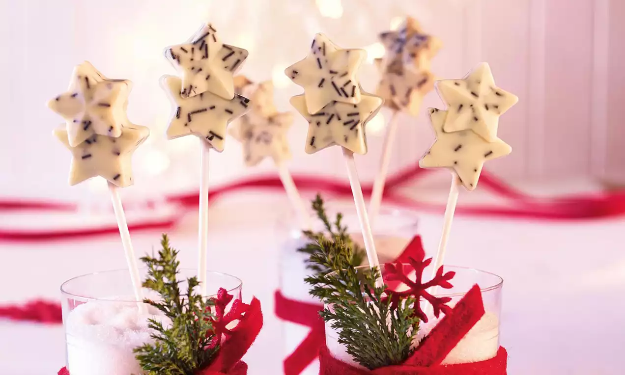 Čokoladne lizalice u obliku zvjezdica od bijele čokolade posute tamnim mrvicama na bijelim štapićima, u čašama s crvenim trakama i zelenim grančicama