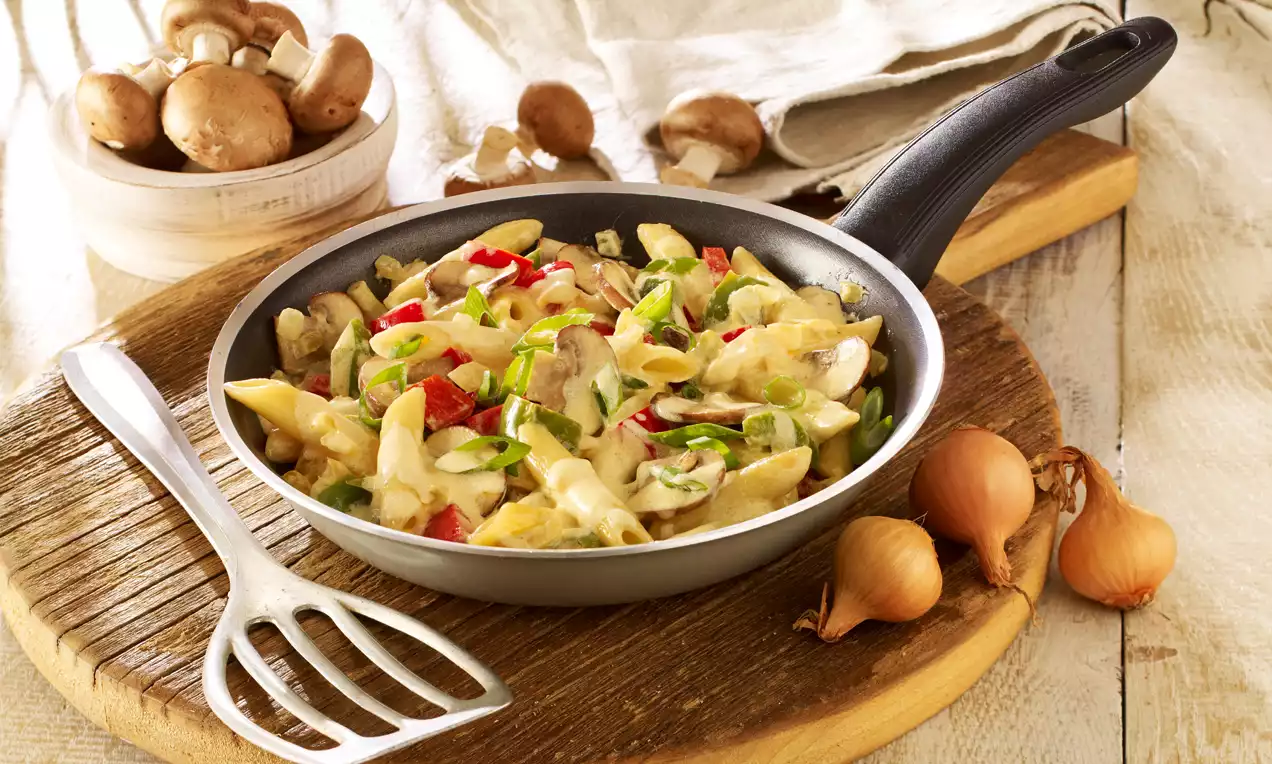 Bunte Nudel-Gemüse-Pfanne in heller Sauce mit Paprikastreifen, Pilzen und Frühlingszwiebeln in einer schwarzen Pfanne auf Holzbrett