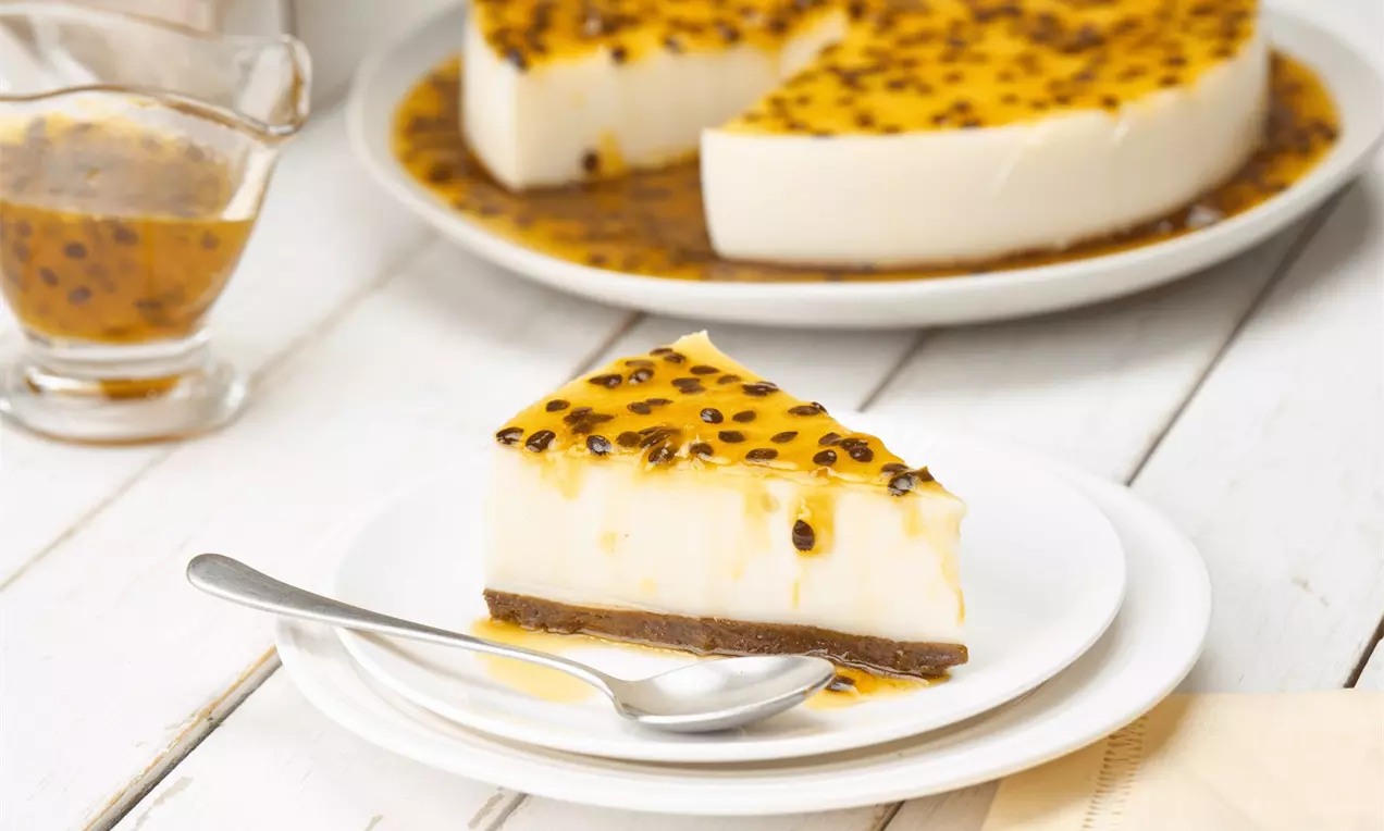 Fatia de torta de maracujá com base marrom, creme branco liso e cobertura dourada com sementes, servida em prato branco com colher prata