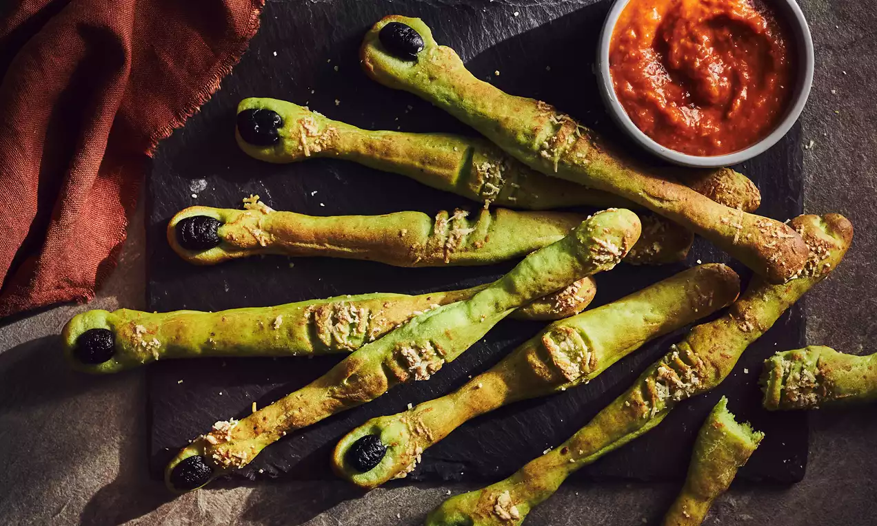Green breadsticks shaped as gnarly fingers with black olive nails, wrinkled knuckles, sprinkled with cheese, served on a slate with red dip