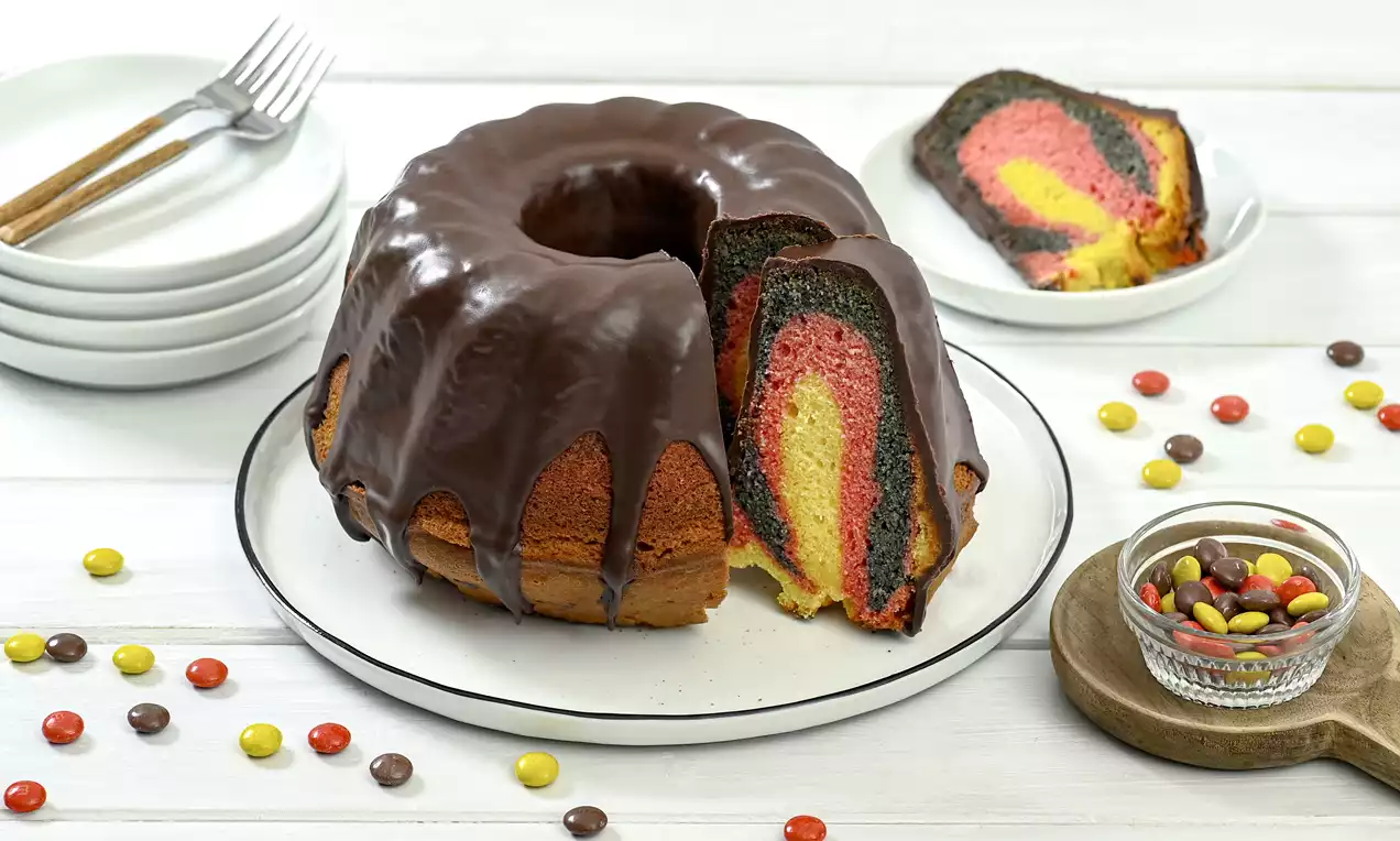 Ringförmiger Kuchen mit glänzender Schokoglasur, farbigem Muster aus Schwarz, Rot und Gelb, auf weißem Teller mit bunter Süßigkeitendekoration
