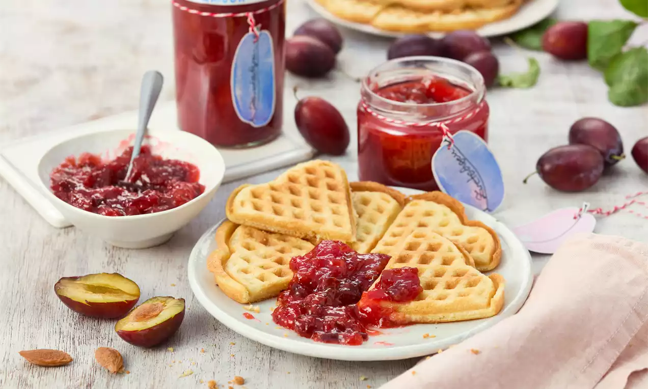 Herzförmige goldene Waffeln auf einem weißen Teller, garniert mit tiefroter, glänzender Pflaumengrütze, daneben frische Pflaumen und Gläser