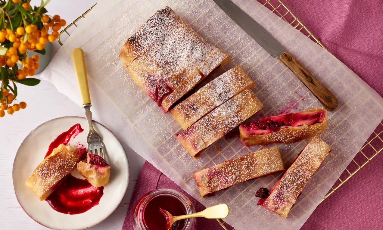 Goldbrauner Strudel mit roter Beerenfüllung und Puderzucker bestäubt, angeschnitten auf Backpapier neben Teller mit Strudelstück und roter Sauce