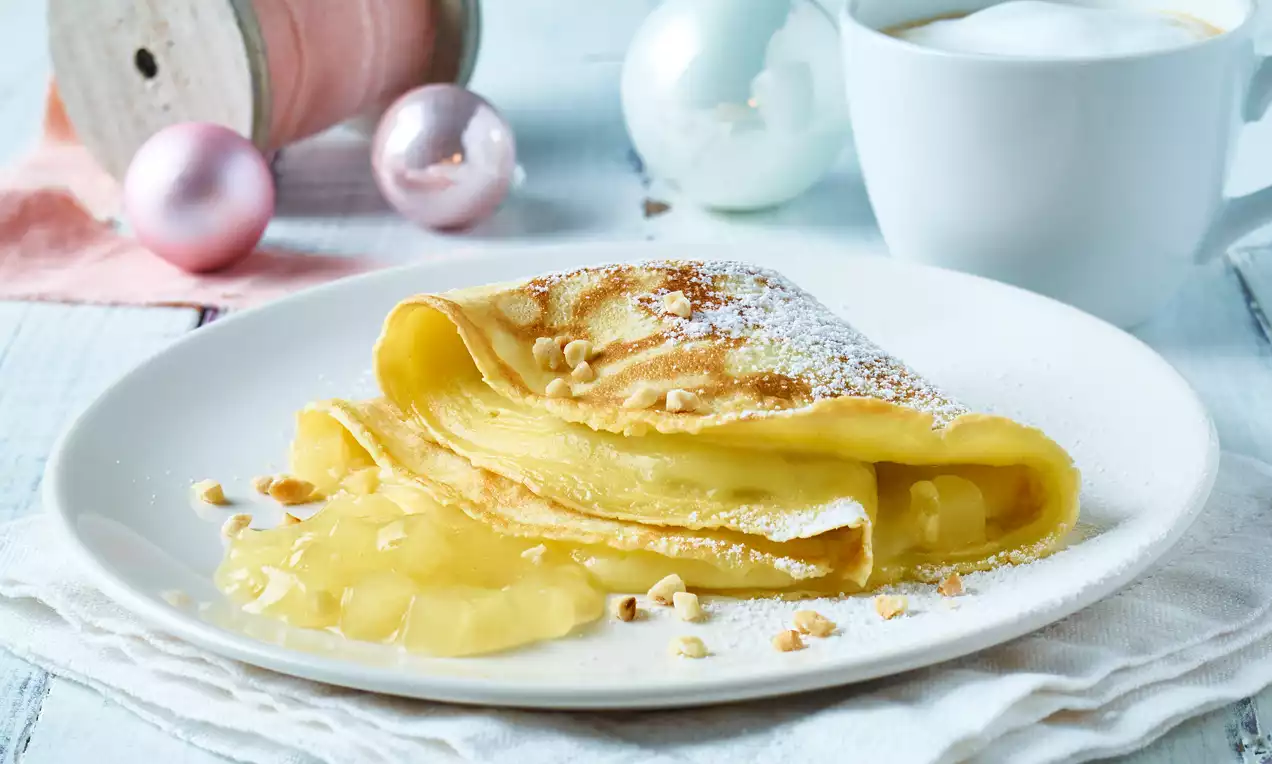Crêpes dorées pliées en triangle, garnies de sauce claire, saupoudrées de sucre glace et de noisettes, sur une assiette blanche