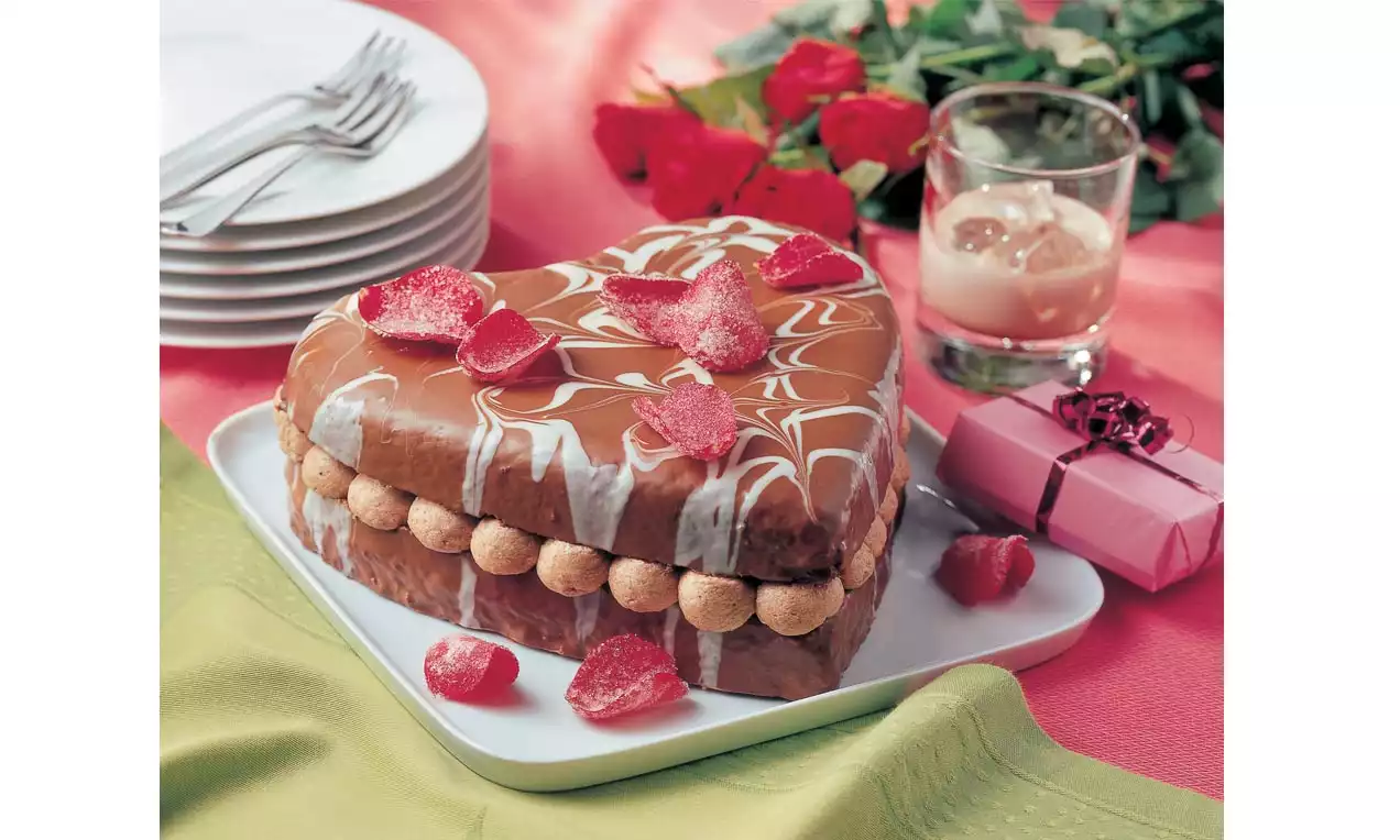 Herzförmige Torte mit glatter Karamellglasur, weißem Zuckermuster, rosa Deko-Herzen und Sahnetupfen am Rand auf weißer Platte