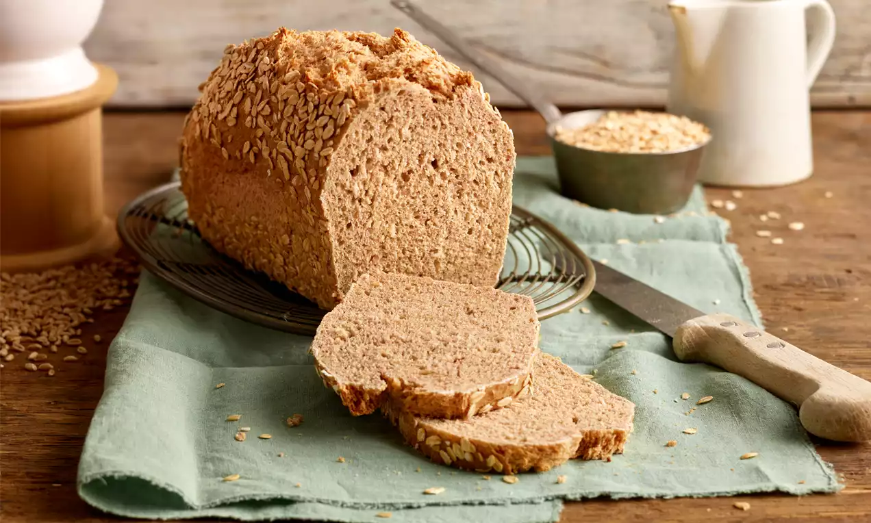 Kastanienbraunes Haferflockenbrot mit knuspriger Kruste, bedeckt mit Haferflocken, auf Holztisch mit grüner Serviette und Brotmesser arrangiert