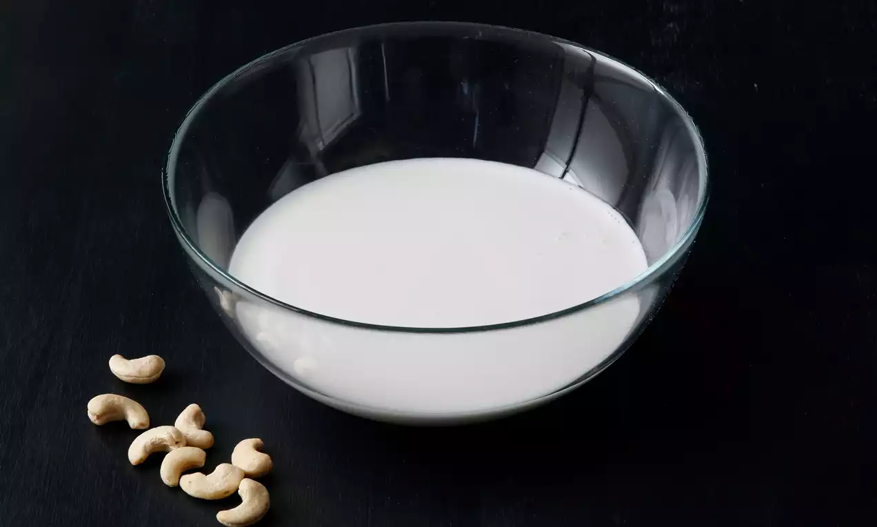 Şeffaf cam bir kasede açık beyaz renkli pürüzsüz kaju sütü, yanında koyu zemine serpiştirilmiş birkaç bütün kaju fıstığı