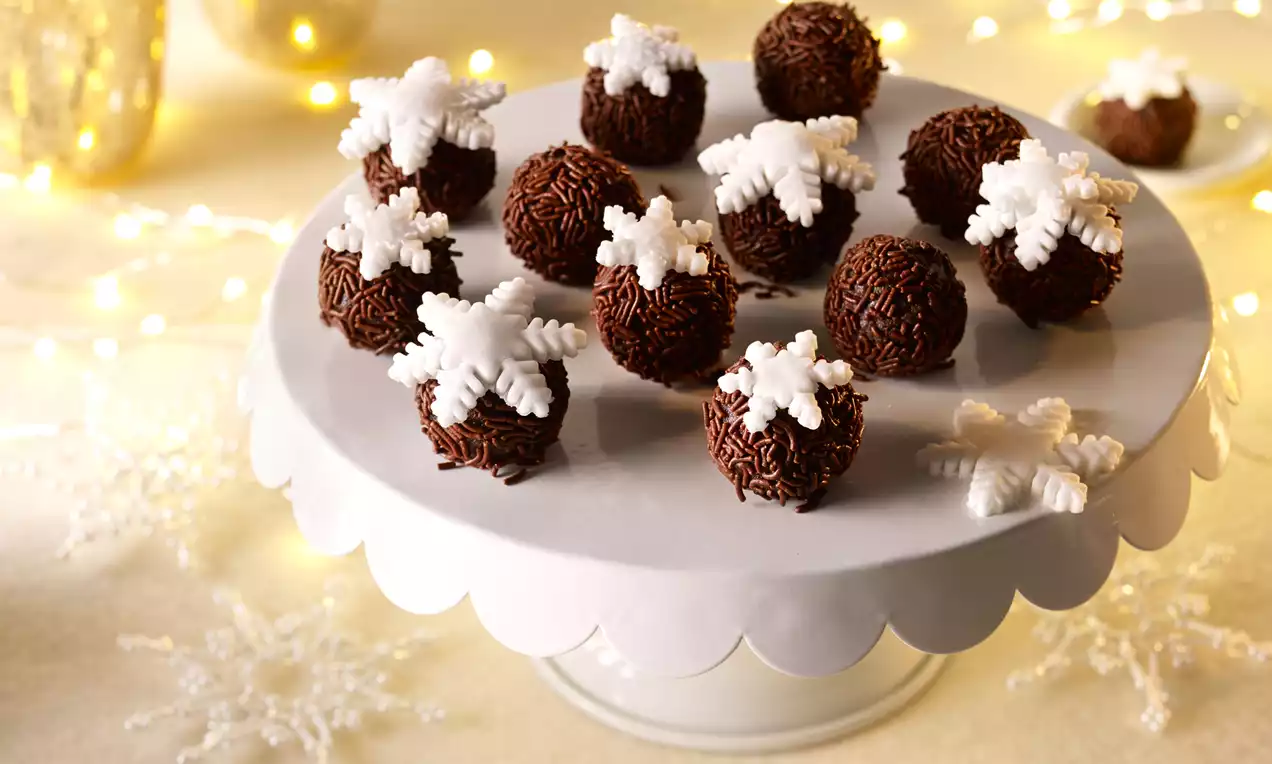 Czekoladowe kulki z ciemną, ziarnistą posypką, udekorowane białymi śnieżynkami na białej paterze z falistym brzegiem w świetle lampek