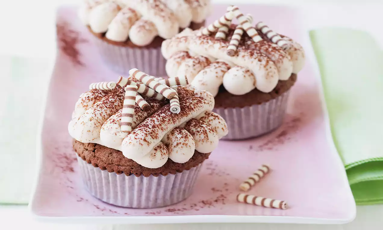 Schokoladen-Muffins mit weißer Cremehaube, bestäubt mit Kakao und garniert mit gestreiften Schokoröllchen auf rosa Teller drapiert