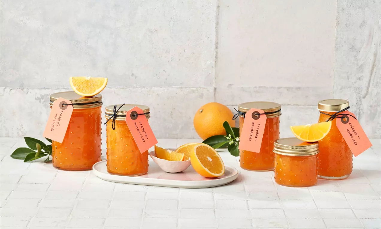 Staklenke narančastog džema s metalnim poklopcima, dekorativnim ceduljama, svježim kriškama i plodovima naranče na bijelom pladnju