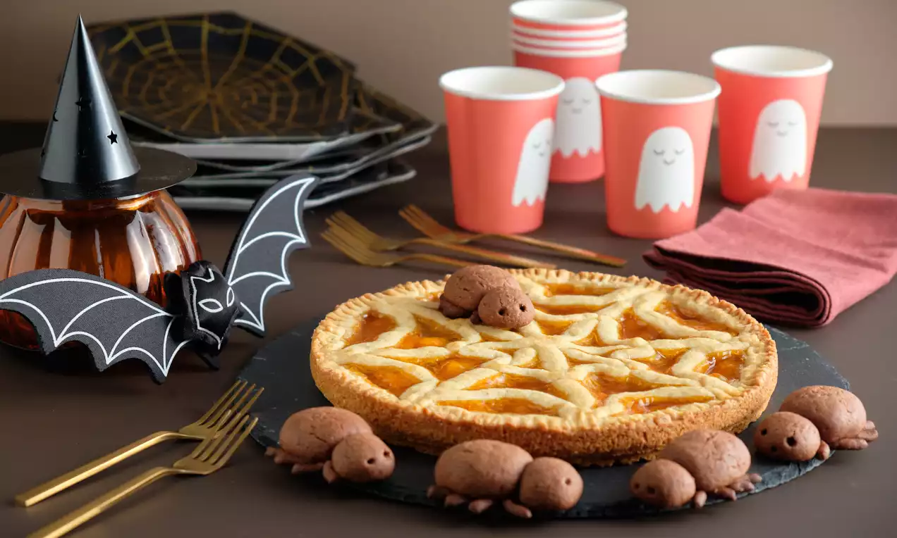 Crostata dorata con ragnatela di pasta intrecciata, ripieno arancione lucido e decorata con dolci ragnetti marroni su piatto nero