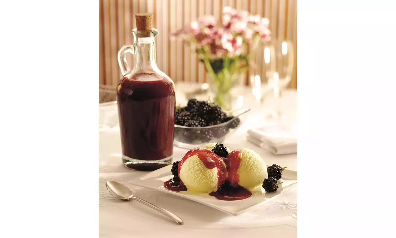 Vanilková zmrzlina s hladkým povrchom, poliata tmavočerveným ovocným toppingom, ozdobená čiernymi lesklými černicami na bielom tanieri