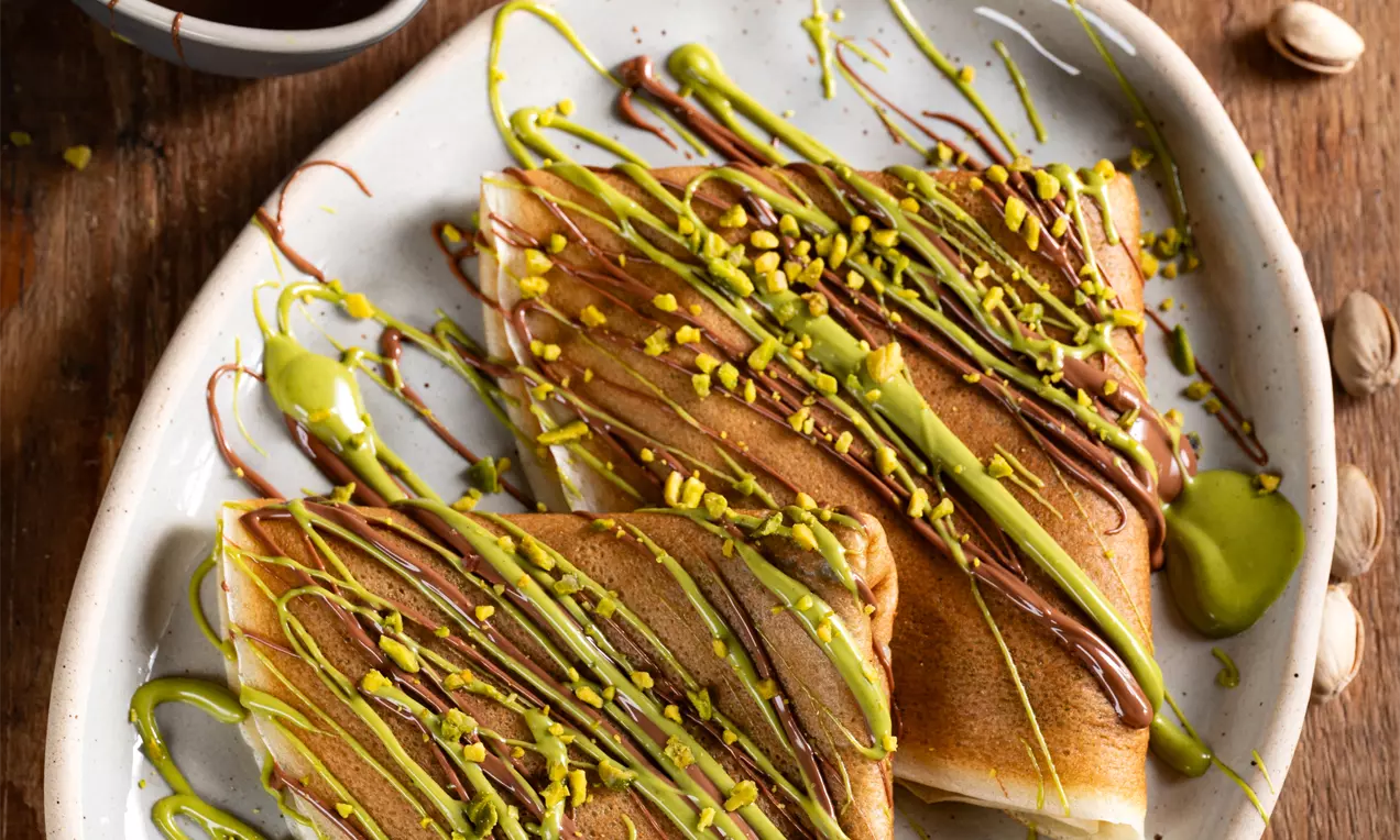 Svijetlo smeđe palačinke trokutastog oblika, prošarane zelenim i smeđim preljevom, ukrašene mljevenim pistacijama na bijelom tanjuru