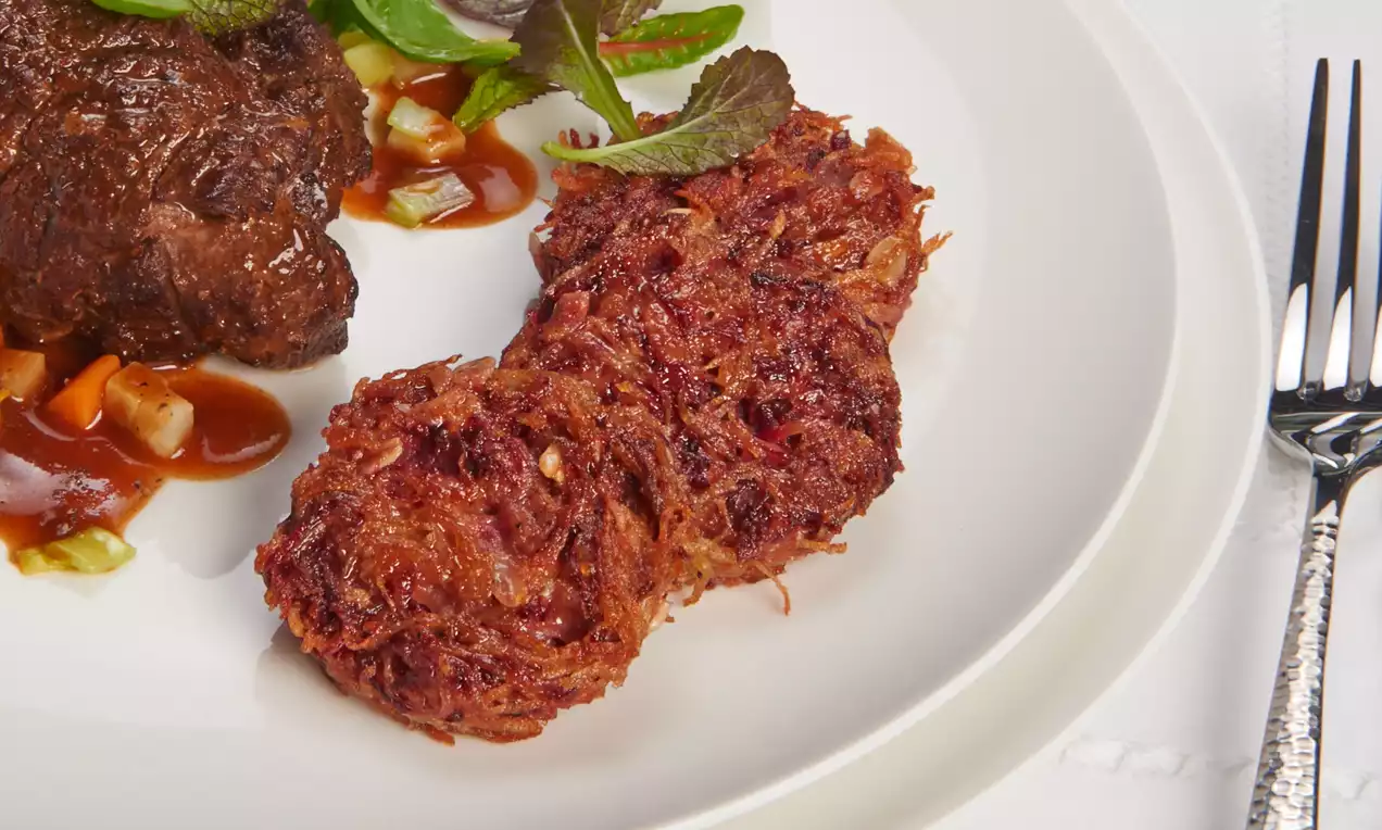 Rote Bete Rösti in länglicher Form, goldbraun-rot gebräunt mit knuspriger Textur, auf einem weißen Teller neben dunklem Braten und Sauce