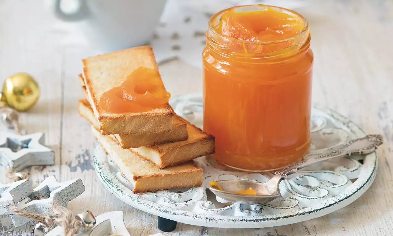 Džem jasne oranžovej farby v sklenenom pohári vedľa kôpok zlatistých toastov, na svetlom tanieri s kovovou lyžičkou