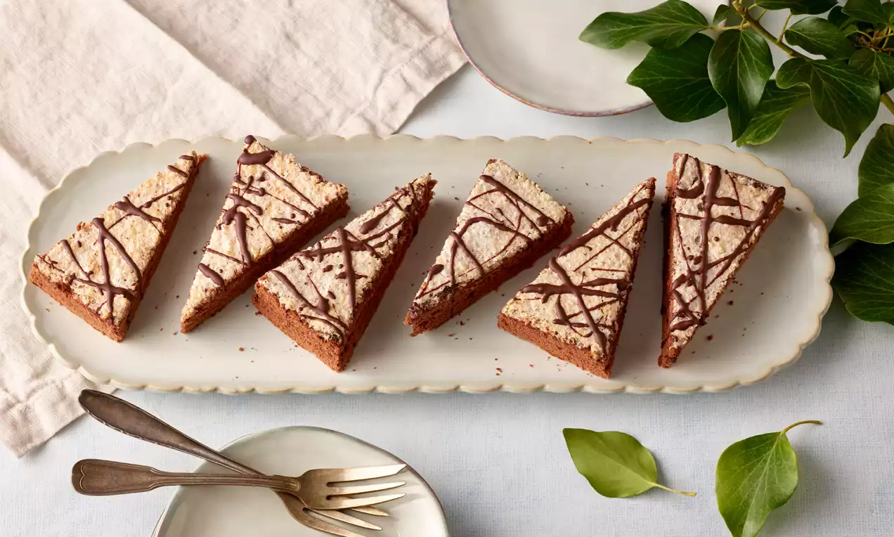 Dreieckige Haselnussschnitten mit heller Cremeschicht und dunkler Schokoladendekoration auf länglicher Platte mit grünen Blättern und Besteck