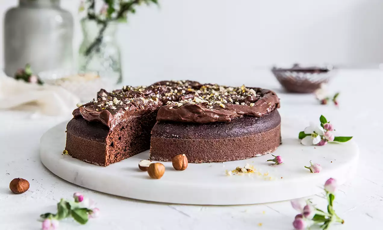 Tmavě hnědý kulatý dort s vláčnou čokoládovou polevou, posypaný sekanými oříšky, na mramorovém podnosu zdobeném kvítky a lískovými ořechy