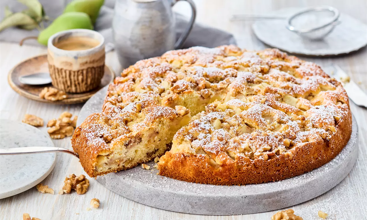 Altın kahverengi cevizli elmalı kek, üstü pudra şekeri serpilmiş, taş tabak üzerinde dilimlenmiş ve cevizlerle çevrelenmiş