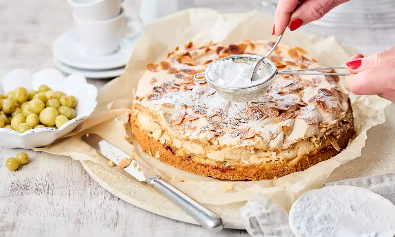 Stachelbeerkuchen mit goldbraunem Teig, luftiger Creme und knuspriger Baiser-Schicht, bestäubt mit Puderzucker auf Backpapier angerichtet