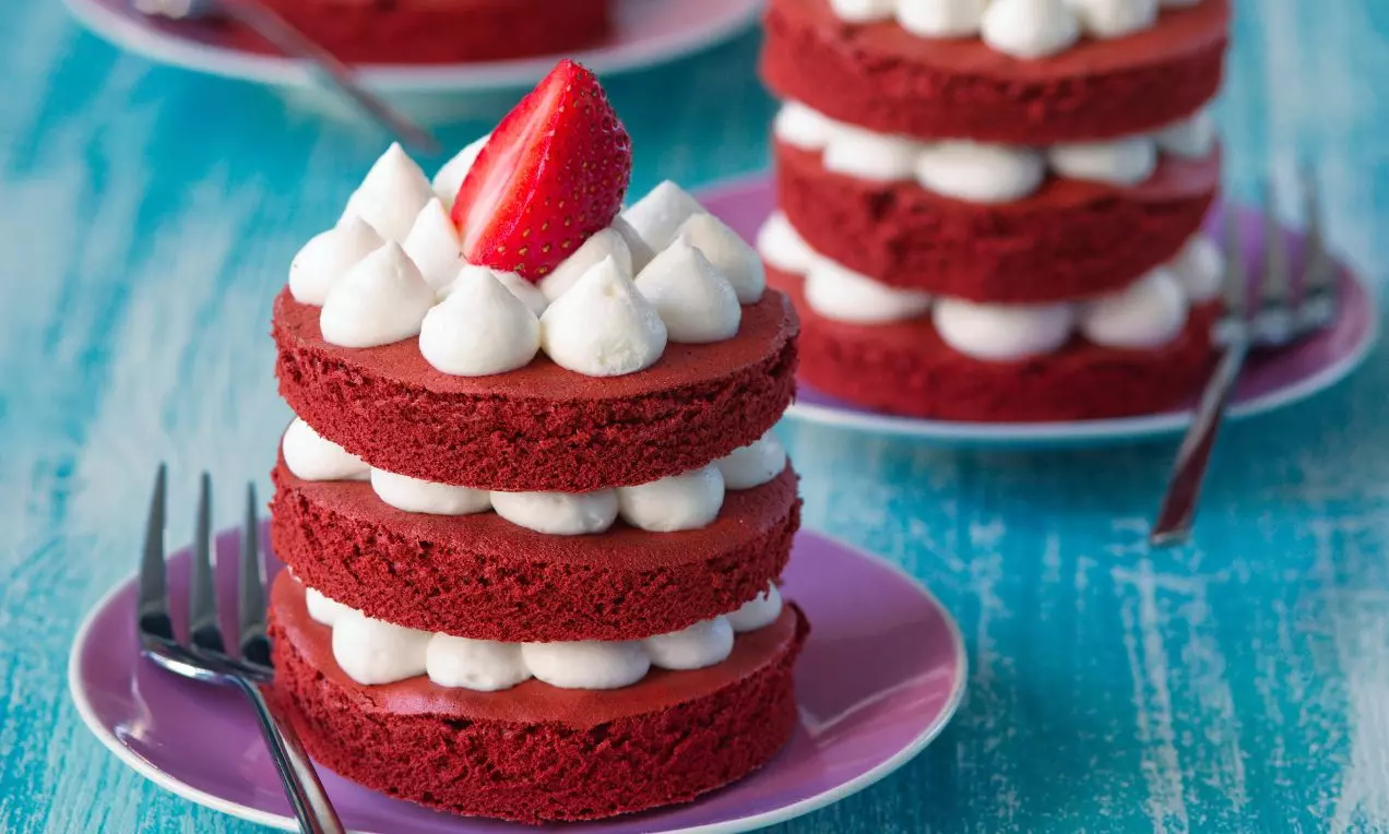 Mini tortička s vrstvami sýto červeného korpusu, medzi ktorými sú biele krémové kopčeky, na vrchu čerstvá polovica jahody na fialovom tanieriku