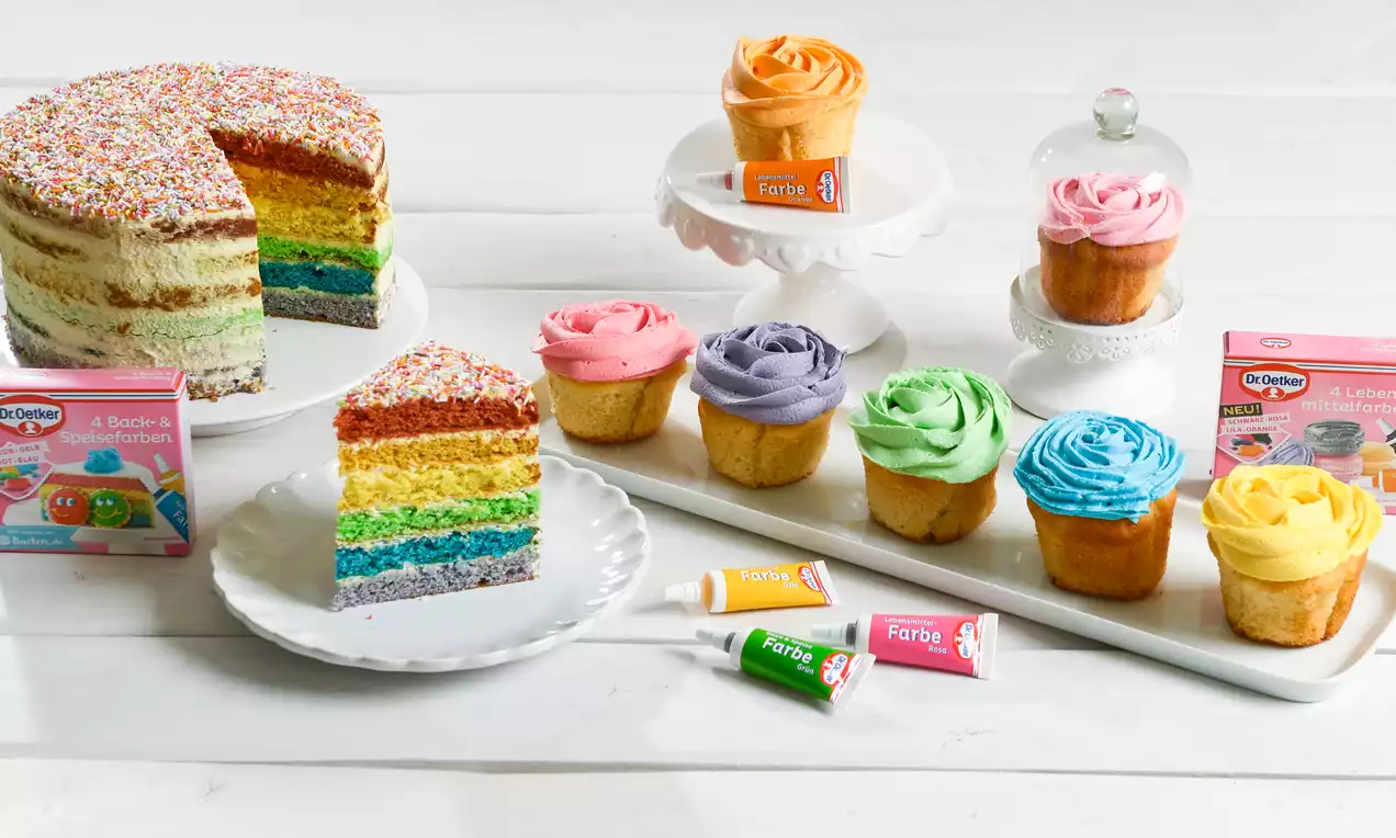 Regenbogenfarbige Schichttorte mit bunter Streuselkruste, daneben Cupcakes mit farbiger Creme auf weißem Geschirr und Lebensmittelfarben-Tuben